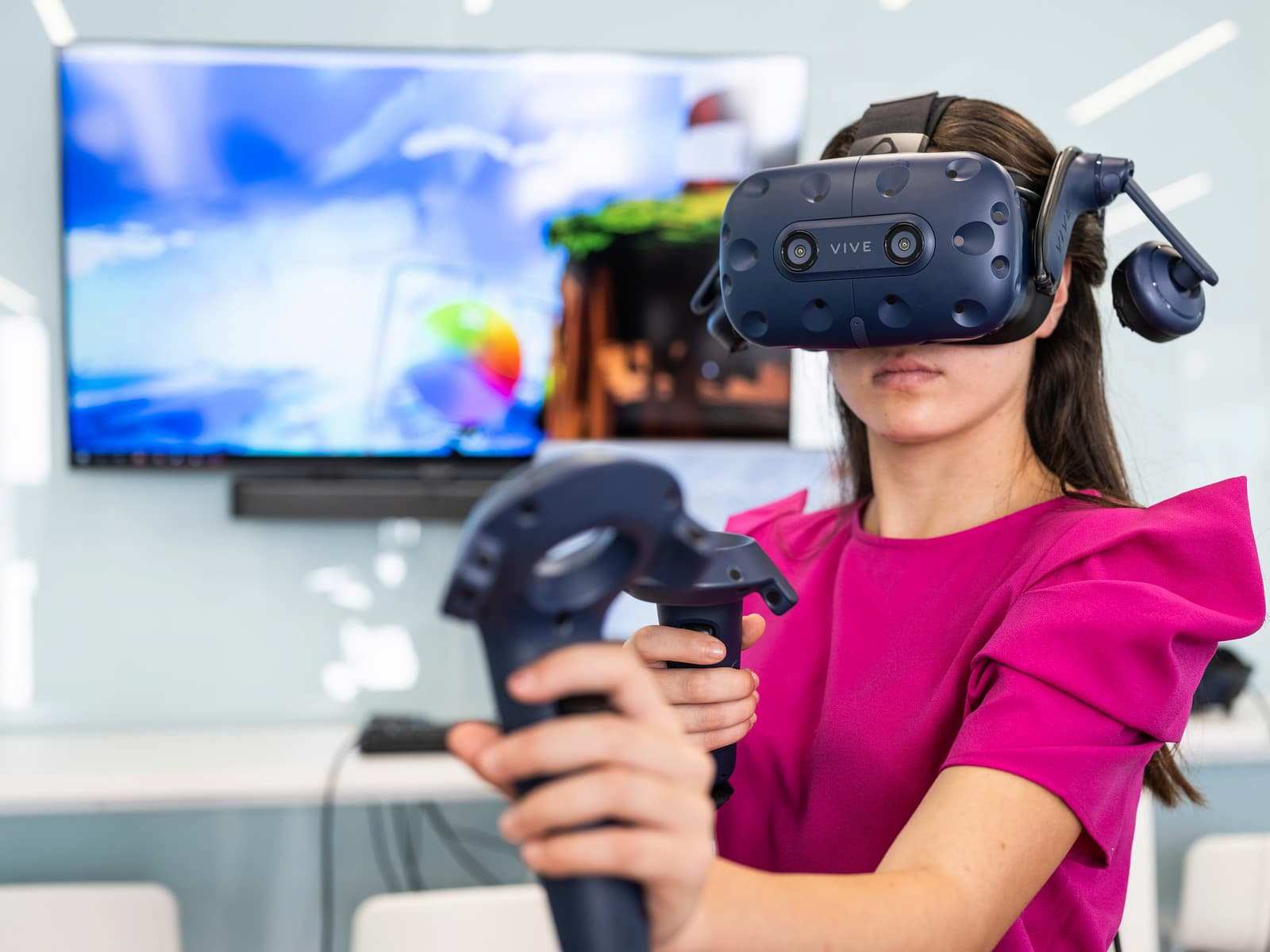 Estudiante con visor y controles de realidad virtual Vive participa en una simulación interactiva frente a una pantalla.